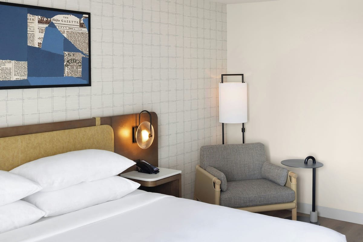 A hotel room with a bed, a chair, a small table, a lamp, and a wall-mounted artwork.