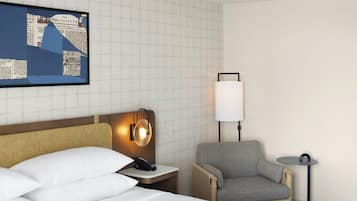 A hotel room with a bed, a chair, a small table, a lamp, and a wall-mounted artwork.