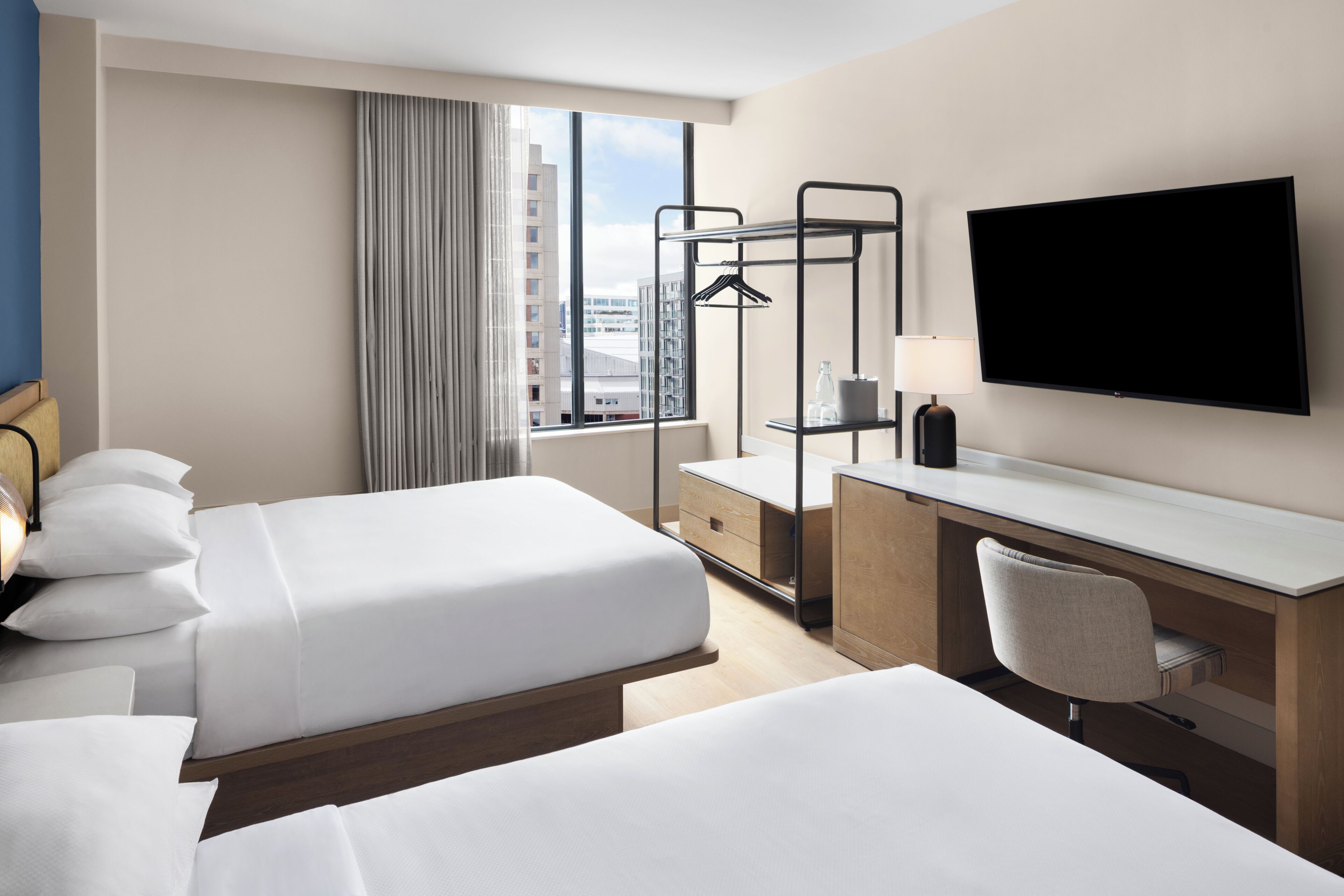 A hotel room with two beds, a desk, a chair, a TV, and a window with a view of buildings.