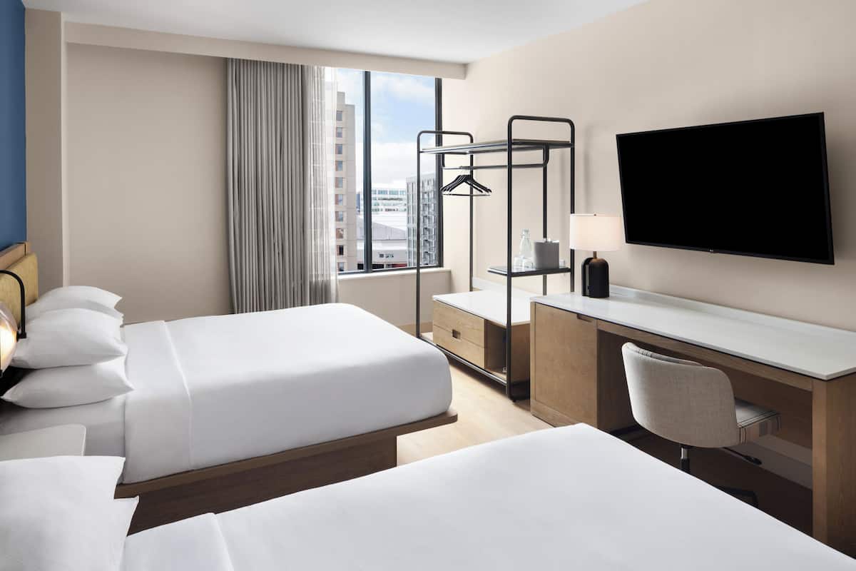 A hotel room with two beds, a desk, a chair, a TV, and a window with a view of buildings.