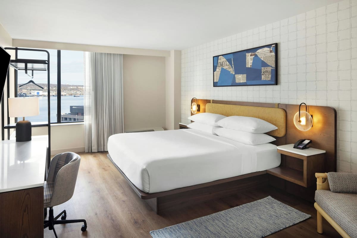 A hotel room with a bed, bedside tables, a desk, a chair, a view of the city, and a cityscape painting on the wall.