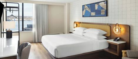 A hotel room with a bed, bedside tables, a desk, a chair, a view of the city, and a cityscape painting on the wall.