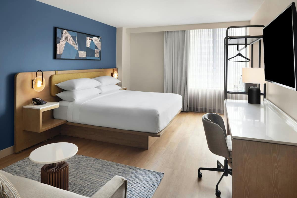 A hotel room with a bed, bedside table, lamp, desk, chair, TV, and a window with curtains.