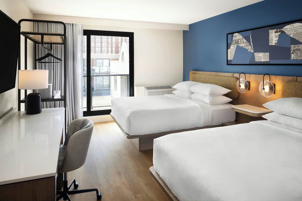 A hotel room with two beds, a desk, a chair, a TV, and a window with a view of buildings.