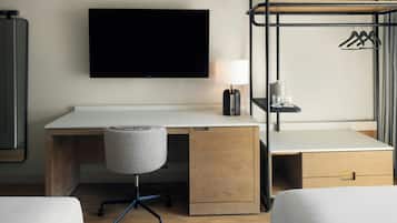 A hotel room with a flat-screen TV, a desk with a lamp, a chair, a bed, and a coat rack.