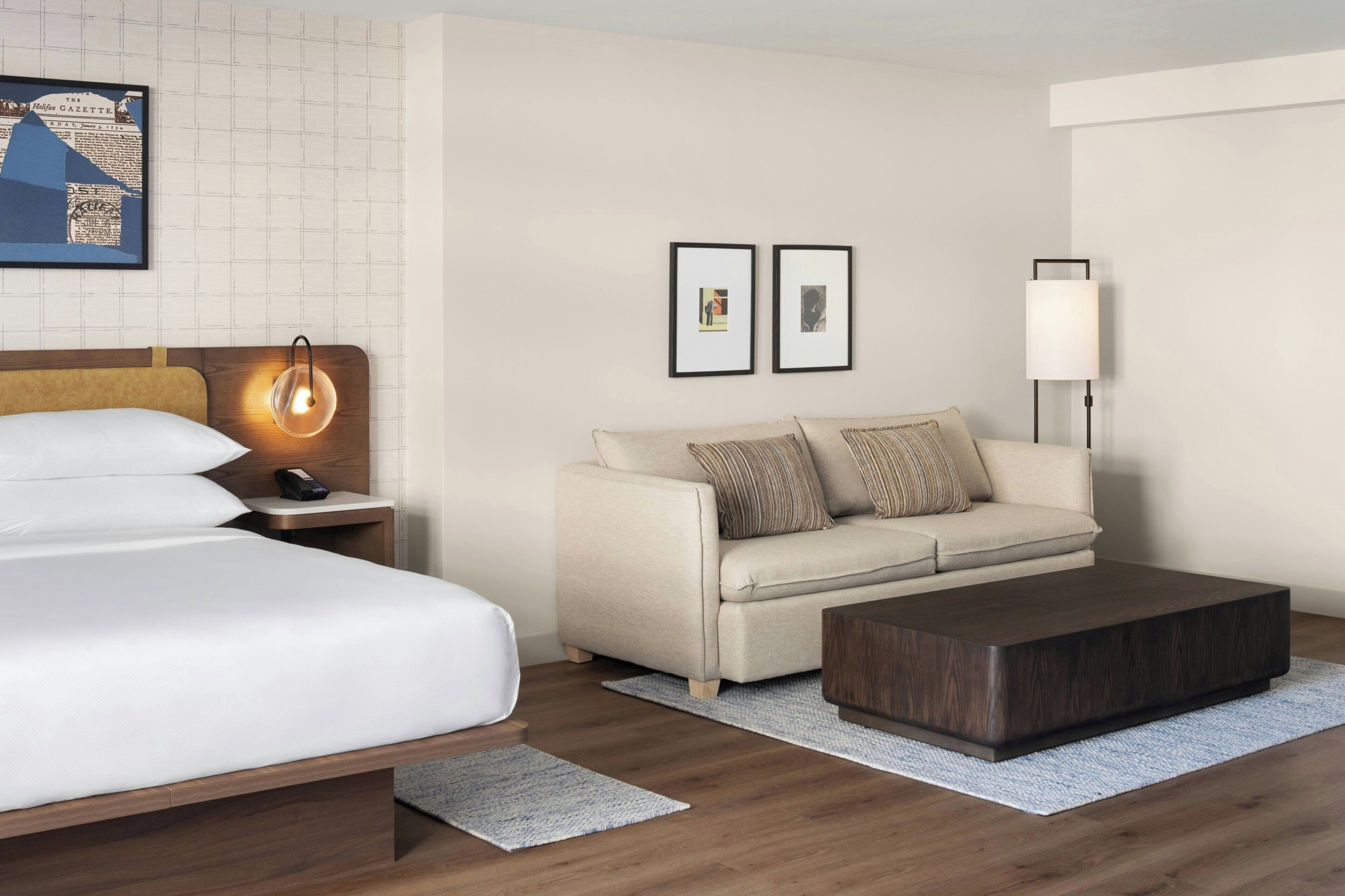 A hotel room with a bed, a sofa, a coffee table, a nightstand, a lamp, and framed pictures on the wall.