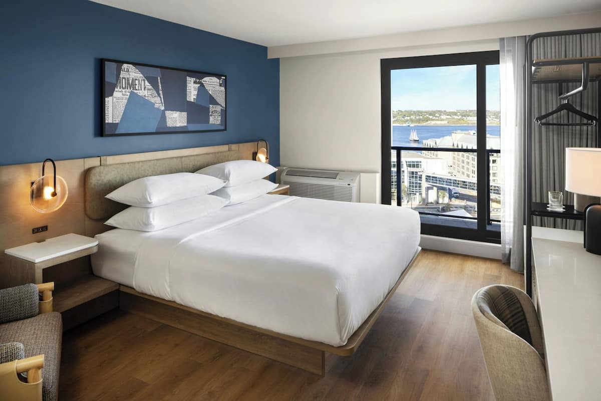 A hotel room with a bed, bedside tables, a chair, a desk, a large window with a city view, and a framed picture on the wall.