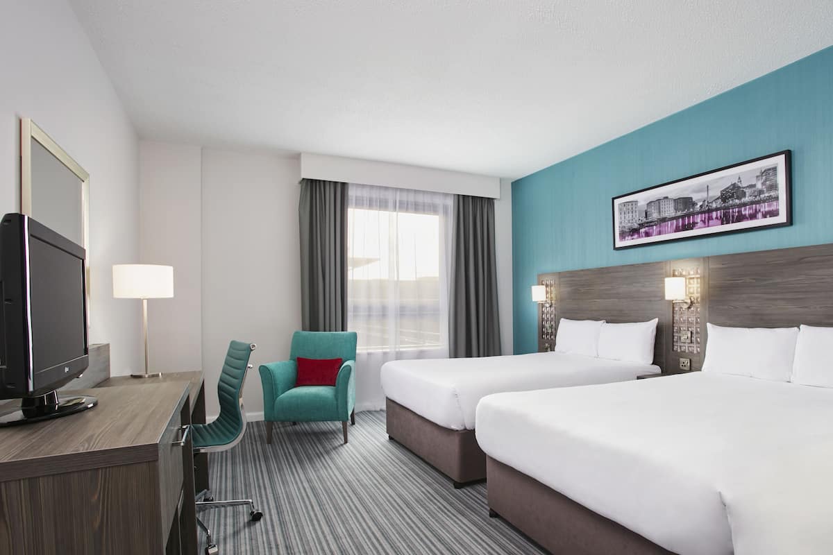 Ein Hotelzimmer mit zwei Betten, einem Schreibtisch mit Fernseher, einem Sessel und einem Fenster mit Vorhängen.