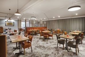 Un comedor de hotel con varias mesas y sillas, un ventanal grande y un elemento decorativo en la pared.