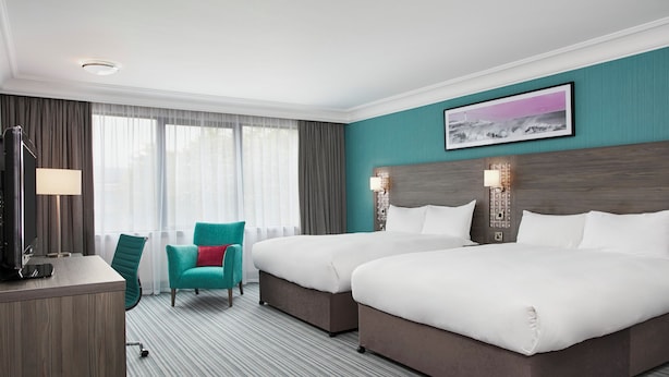 Habitación de hotel con dos camas, un escritorio con lámpara, un sillón color turquesa y un ventanal con cortinas.