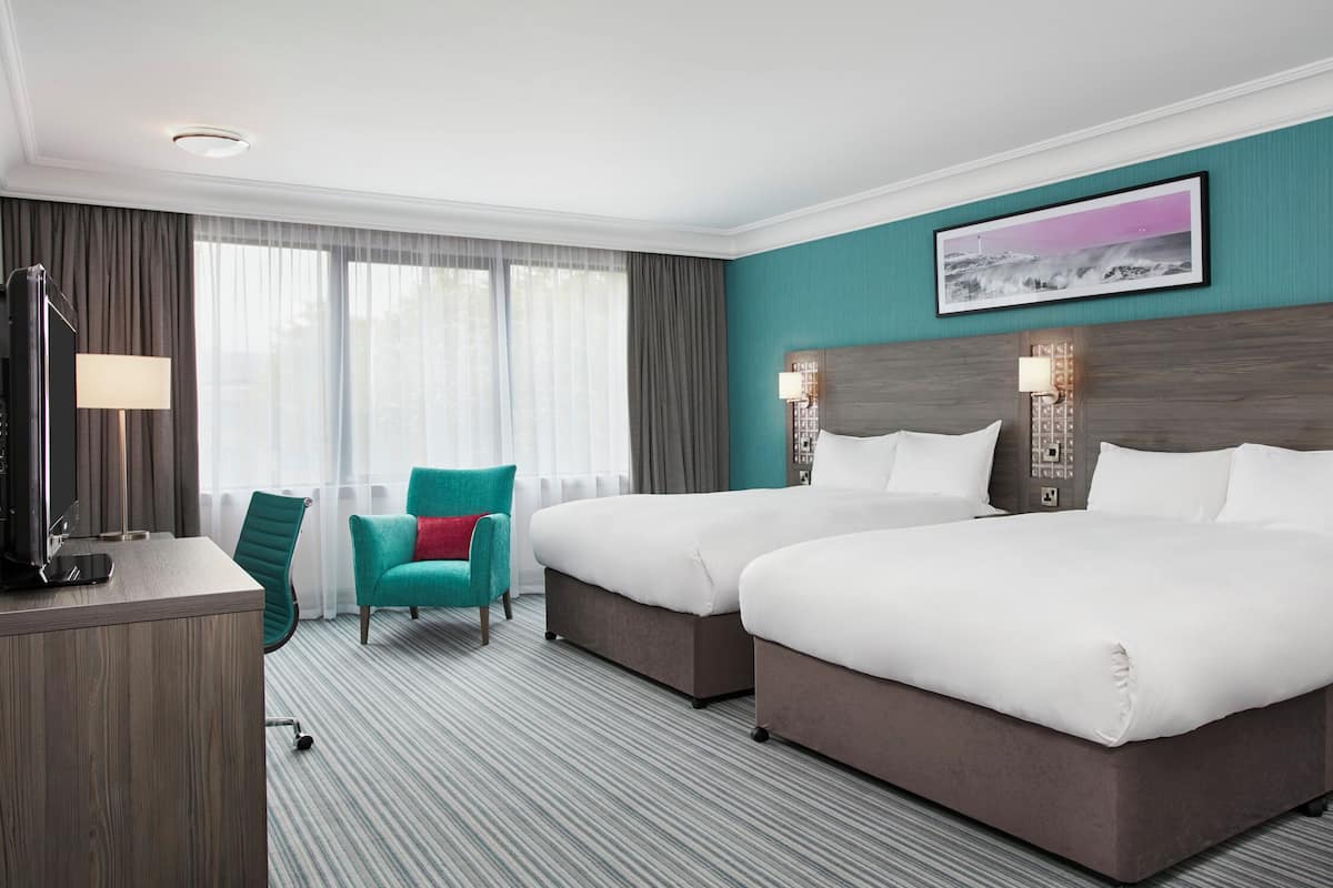 Ein Hotelzimmer mit zwei Betten, einem Schreibtisch mit Lampe, einem blaugrünen Sessel und einem großen Fenster mit Vorhängen.