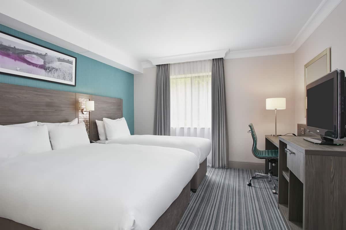 Ein Hotelzimmer mit zwei Betten, einem Schreibtisch mit Computer, einem Fernseher und einem großen Fenster mit Vorhängen.