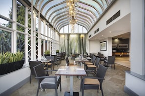 A spacious indoor dining area with high ceilings and large windows, featuring wicker furniture and a modern chandelier.