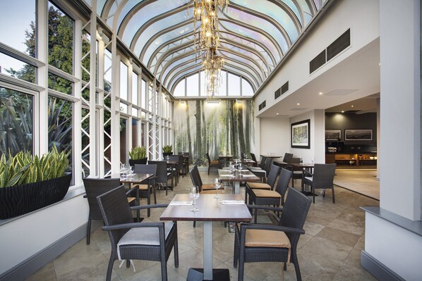 A spacious indoor dining area with high ceilings and large windows, featuring wicker furniture and a modern chandelier.