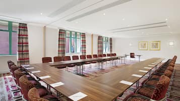 A conference room with a long table and chairs, large windows with curtains, and a carpeted floor.