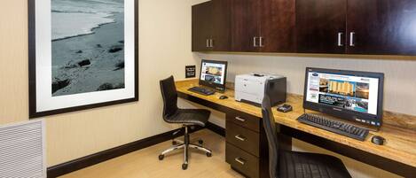 A hotel room with a wooden desk, two computer monitors, a printer, and a framed picture of a beach.