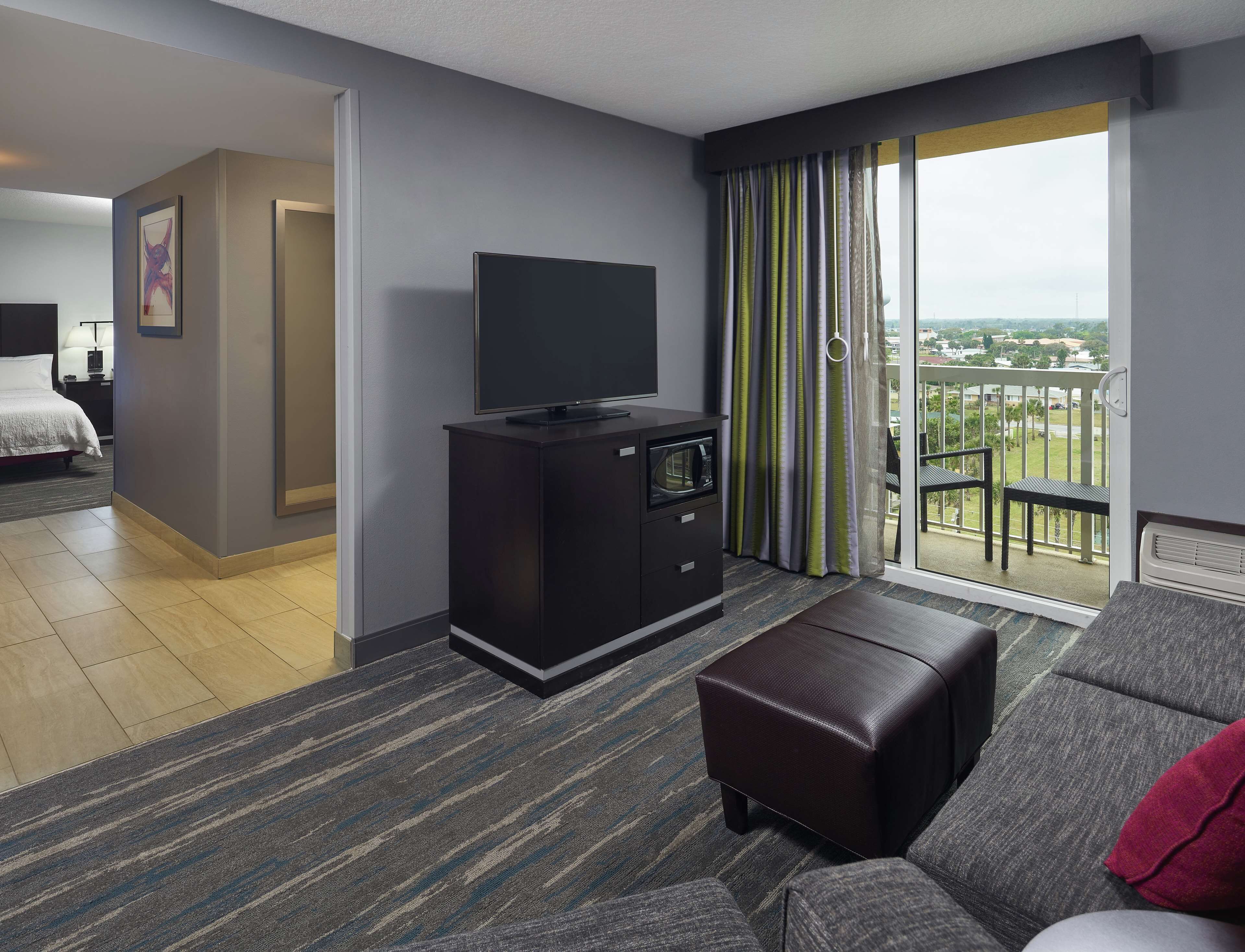A hotel room with a flat-screen TV on a dark wooden cabinet, a gray sofa, a brown ottoman, and a balcony with a view of the city.