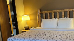 Una habitación de hotel con cama, ventilador de techo, mesita de noche con lámpara y cabecero de madera.