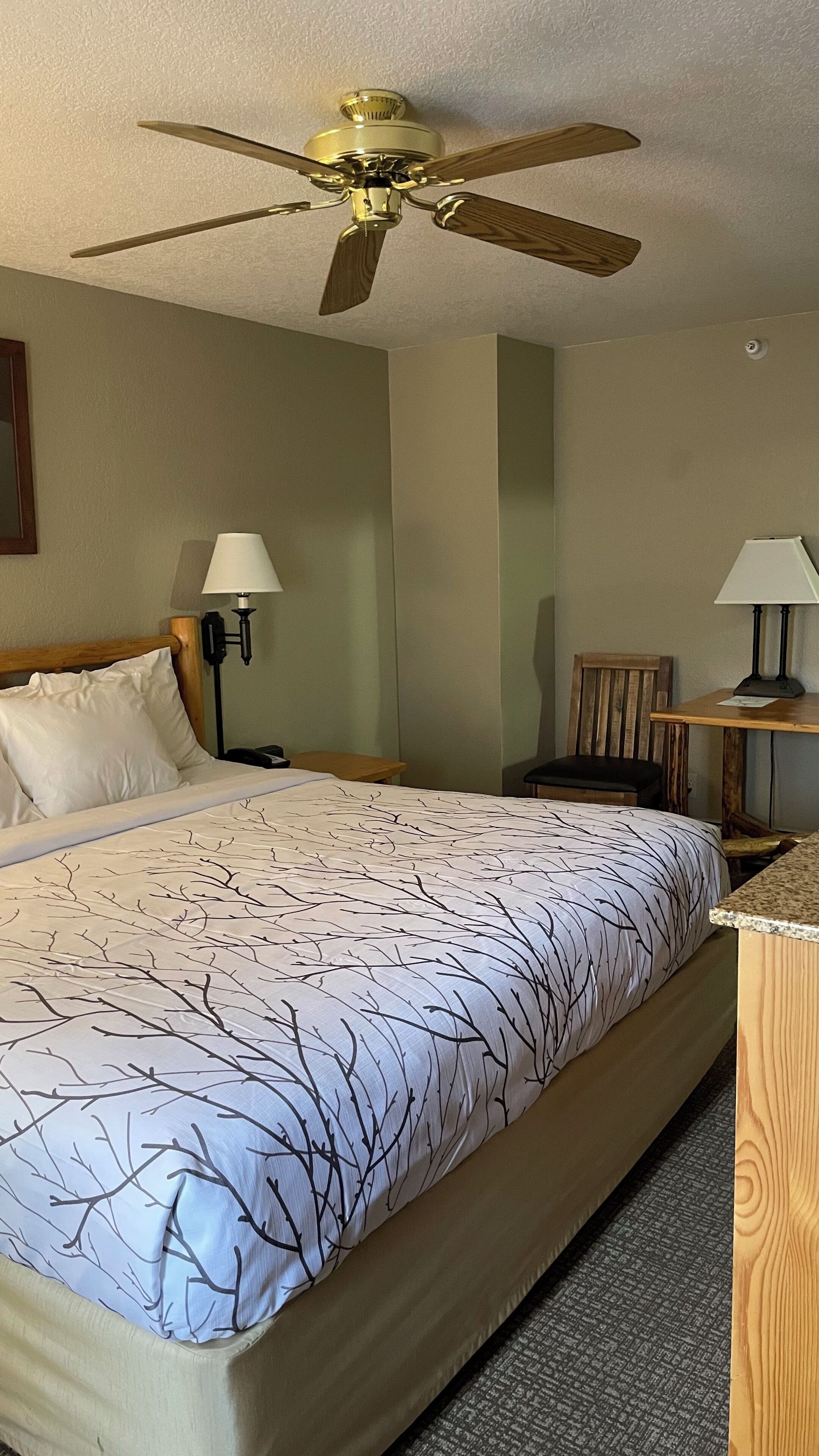 Une chambre d’hôtel comprenant un lit, deux lampes de chevet, un ventilateur de plafond, une table de chevet en bois, une chaise et un bureau avec une lampe.
