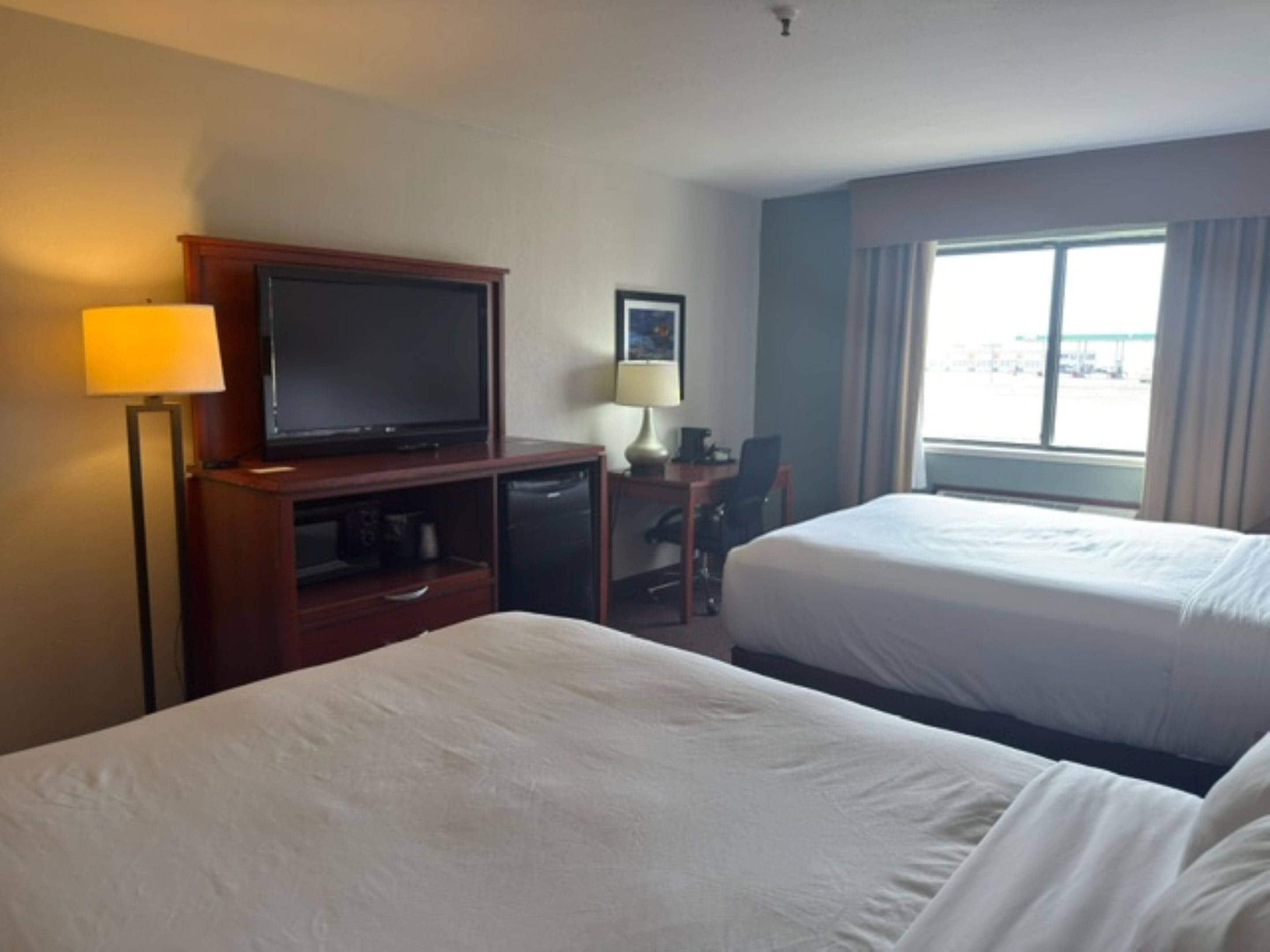 A hotel room with two beds, a desk with a television, a lamp, and a window with a view of the parking lot.