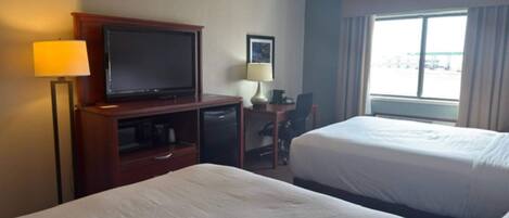 A hotel room with two beds, a desk with a television, a lamp, and a window with a view of the parking lot.