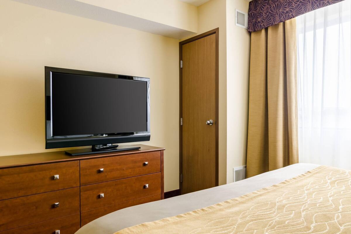A hotel room with a bed, a TV on a wooden dresser, a wooden door, and a window with curtains.