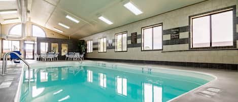 An indoor swimming pool with clear water, a lifeguard on duty sign, and a sunlit ceiling.