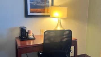 A hotel room with a desk, a chair, a lamp, a framed picture on the wall, and a coffee maker.
