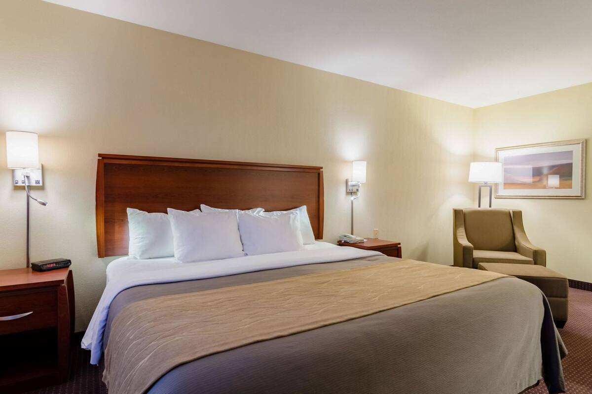 A hotel room with a large bed, a wooden headboard, two bedside tables with lamps, a chair, and a framed picture on the wall.