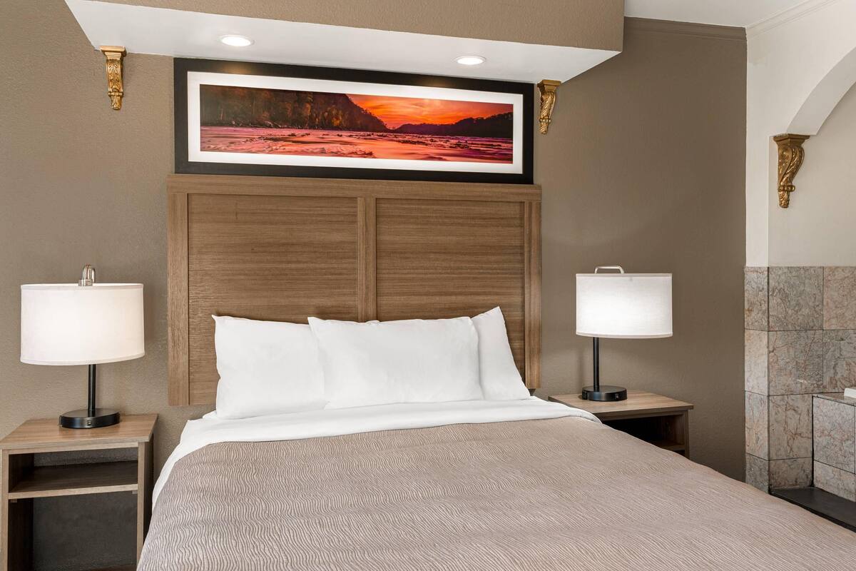 A hotel room with a large bed, two bedside tables with lamps, a wooden headboard, a framed picture above the bed, and a marble-tiled wall.