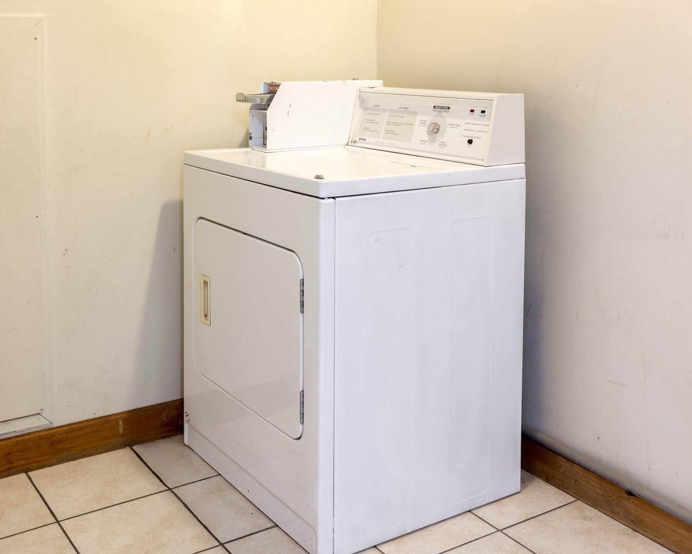 A compact, white front-loading washer and dryer unit in a corner of a room with tiled flooring and wooden trim.