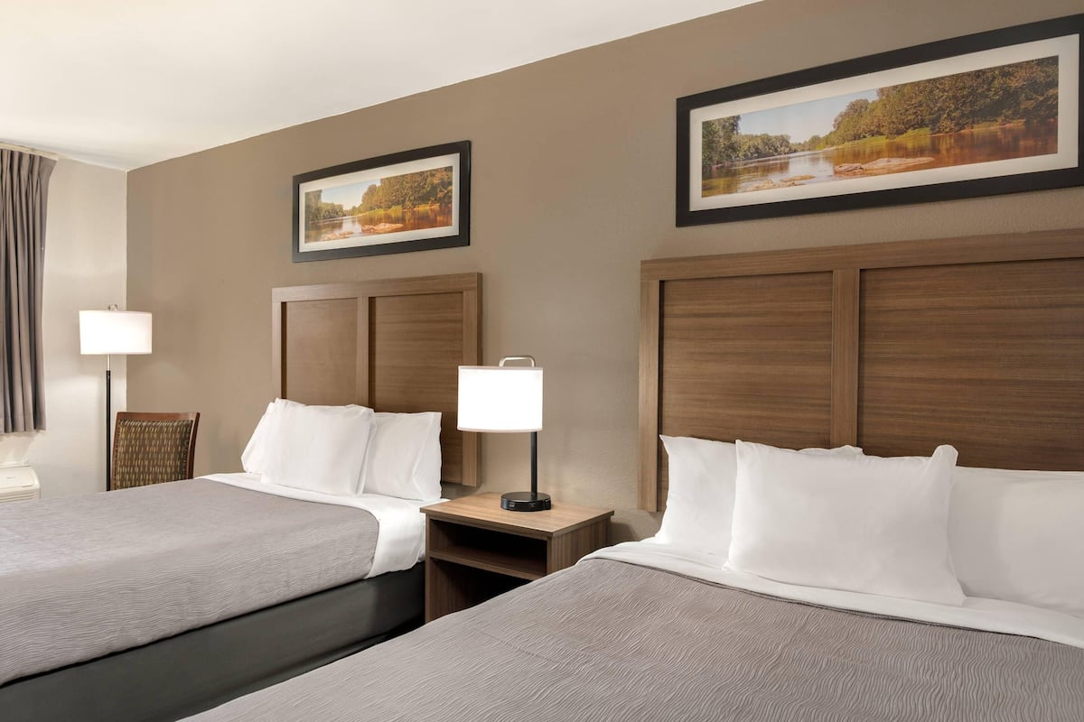 A hotel room with two beds, a wooden headboard, a nightstand with a lamp, and framed pictures on the wall.