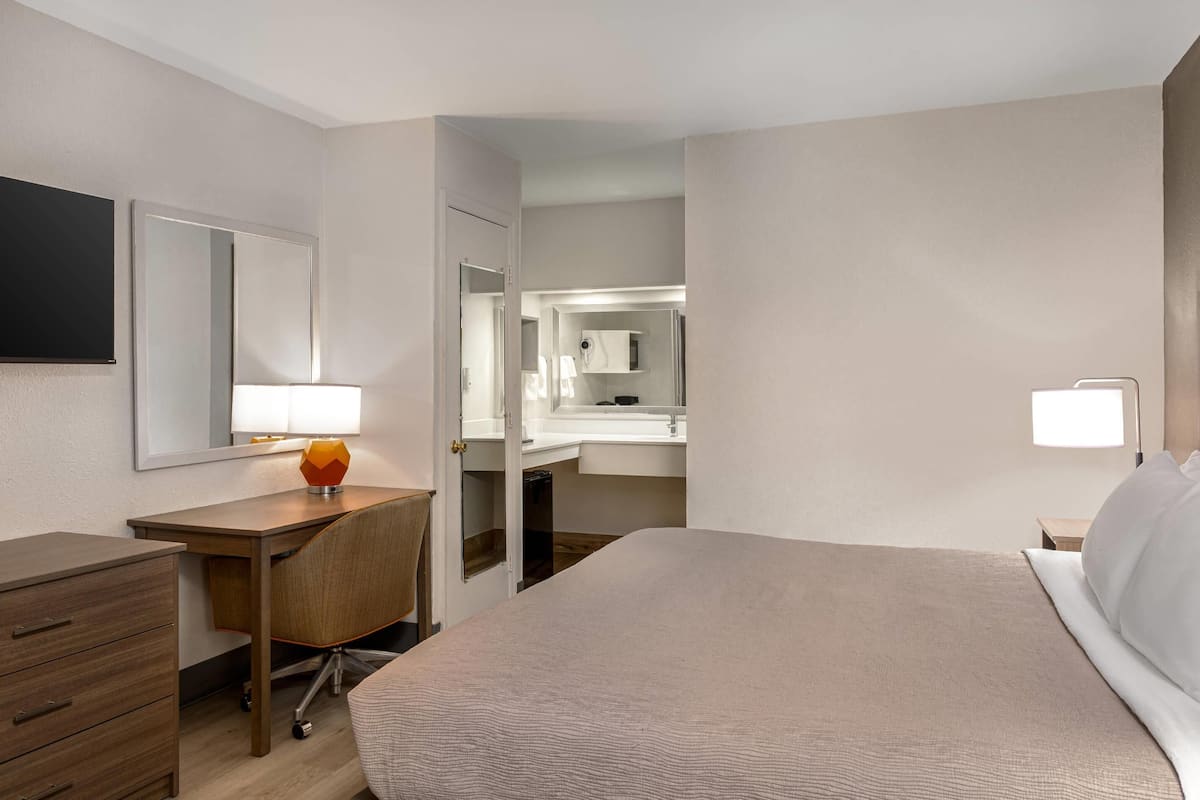 A hotel room with a bed, a desk, a chair, a lamp, a mirror, and a bathroom visible through an open door.