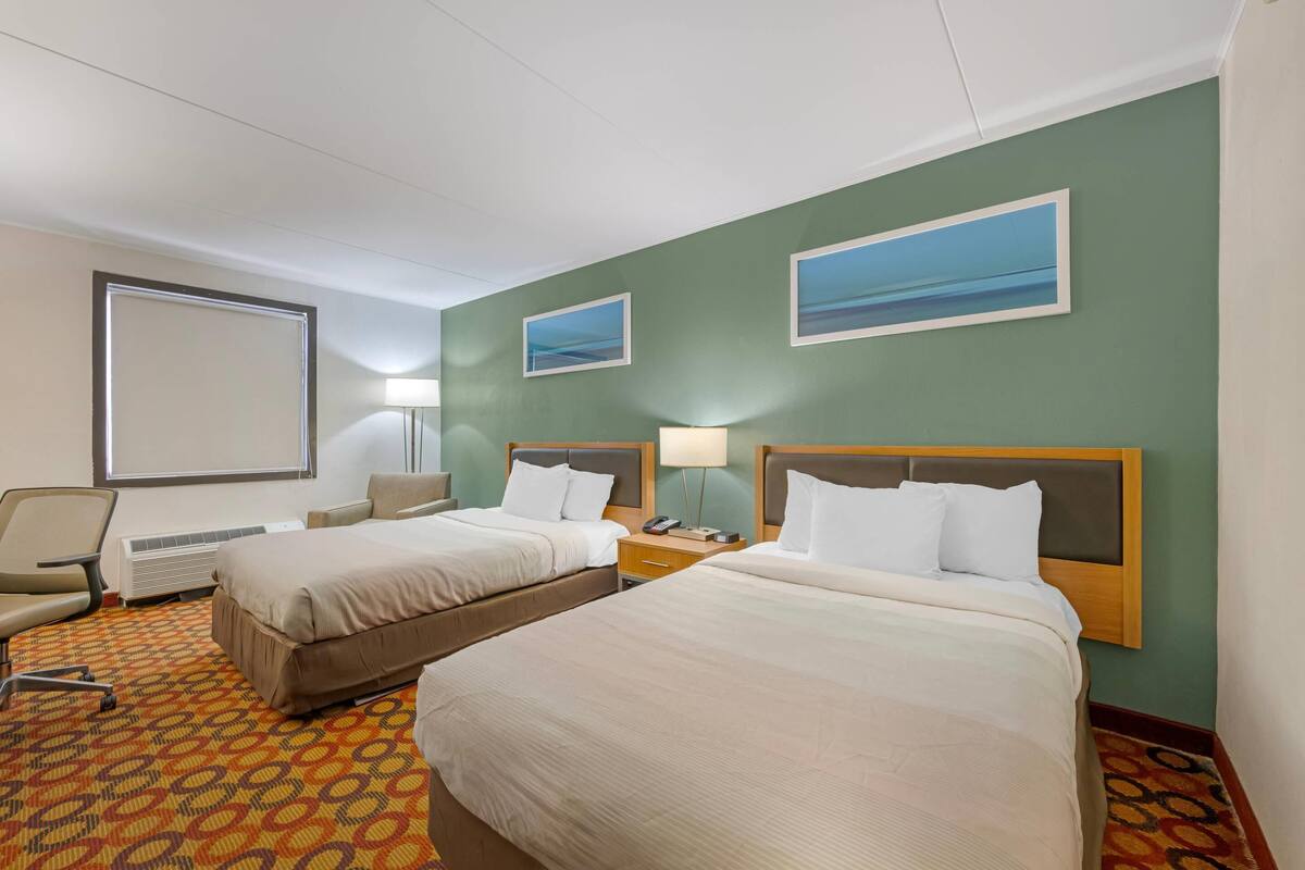 A hotel room with two beds, a desk, a chair, a projector screen, and two wall-mounted lamps.