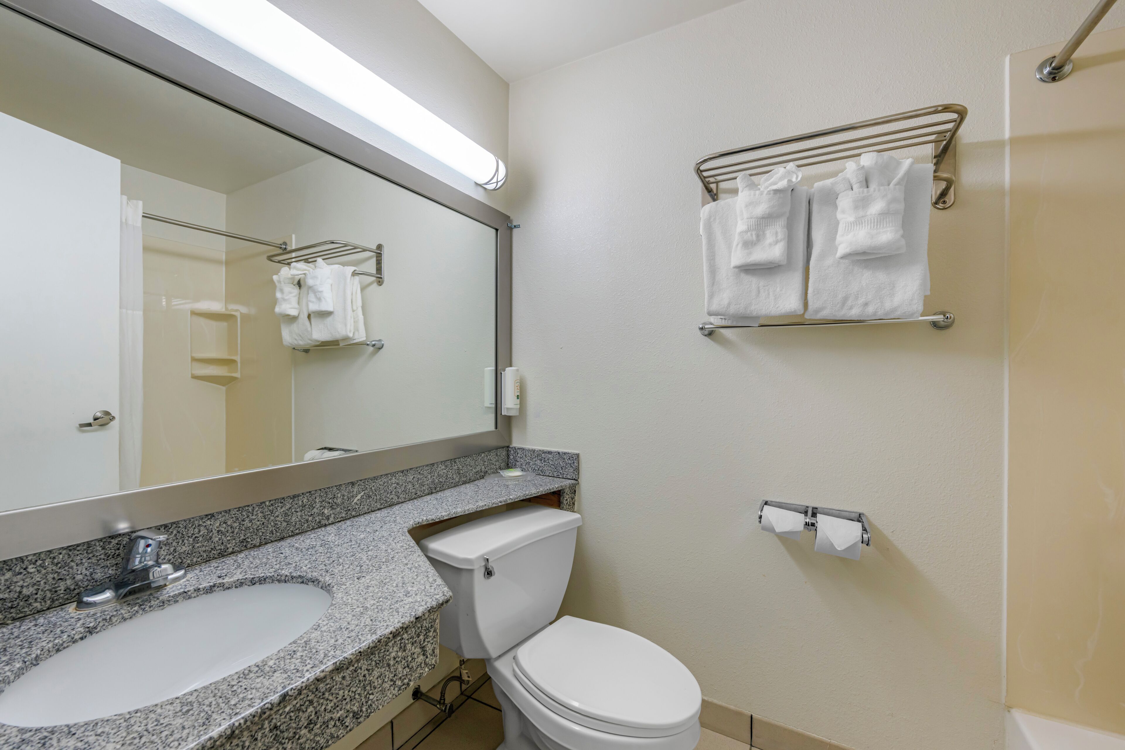 Un baño con lavamanos de encimera de granito, inodoro blanco, toallero con toallas y espejo con luz superior.