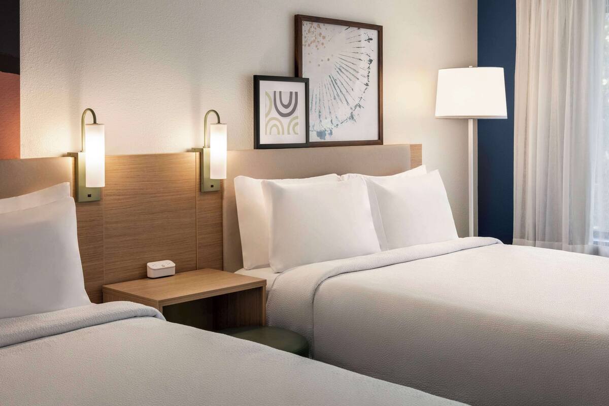 A hotel room with two beds, a bedside table with a lamp, a wall-mounted artwork, and a window with curtains.