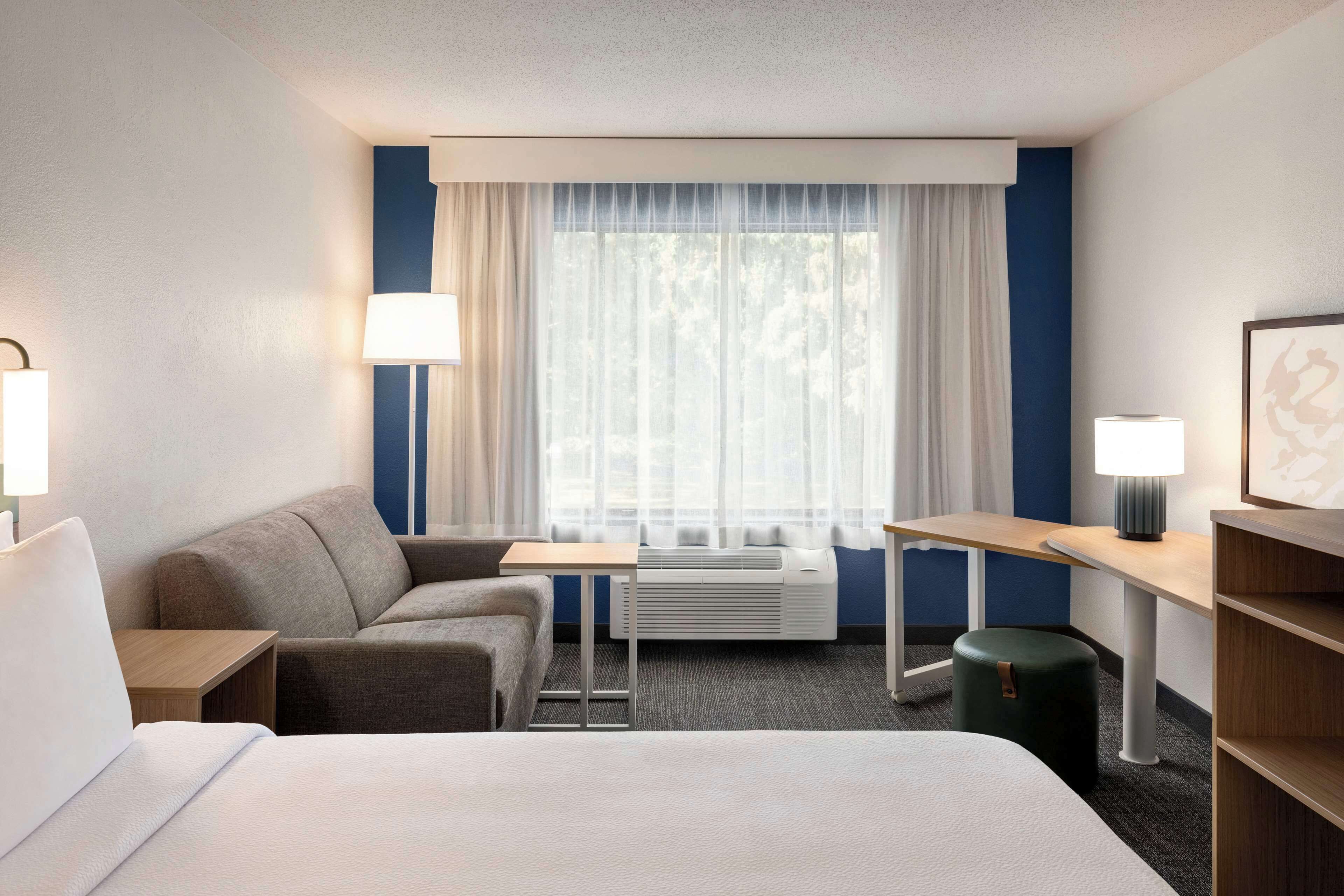 A hotel room with a bed, a sofa, a small table, a lamp, a heater, and a window with curtains.