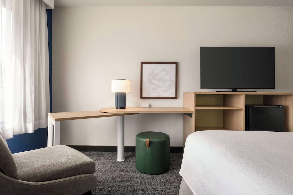A hotel room with a bed, a desk with a lamp, a television, and a small green ottoman.