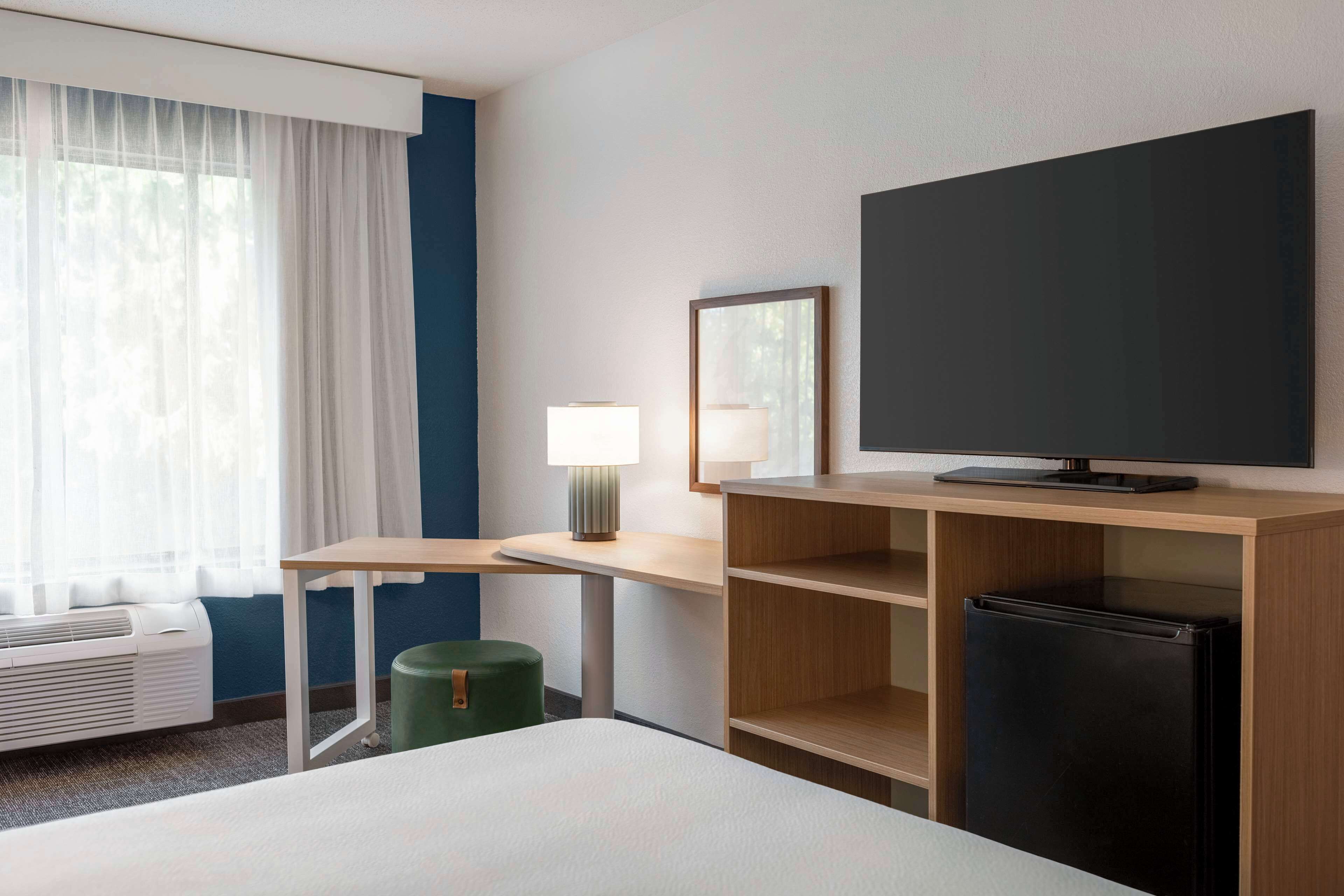 A hotel room with a bed, a desk with a lamp, a television, and a refrigerator.