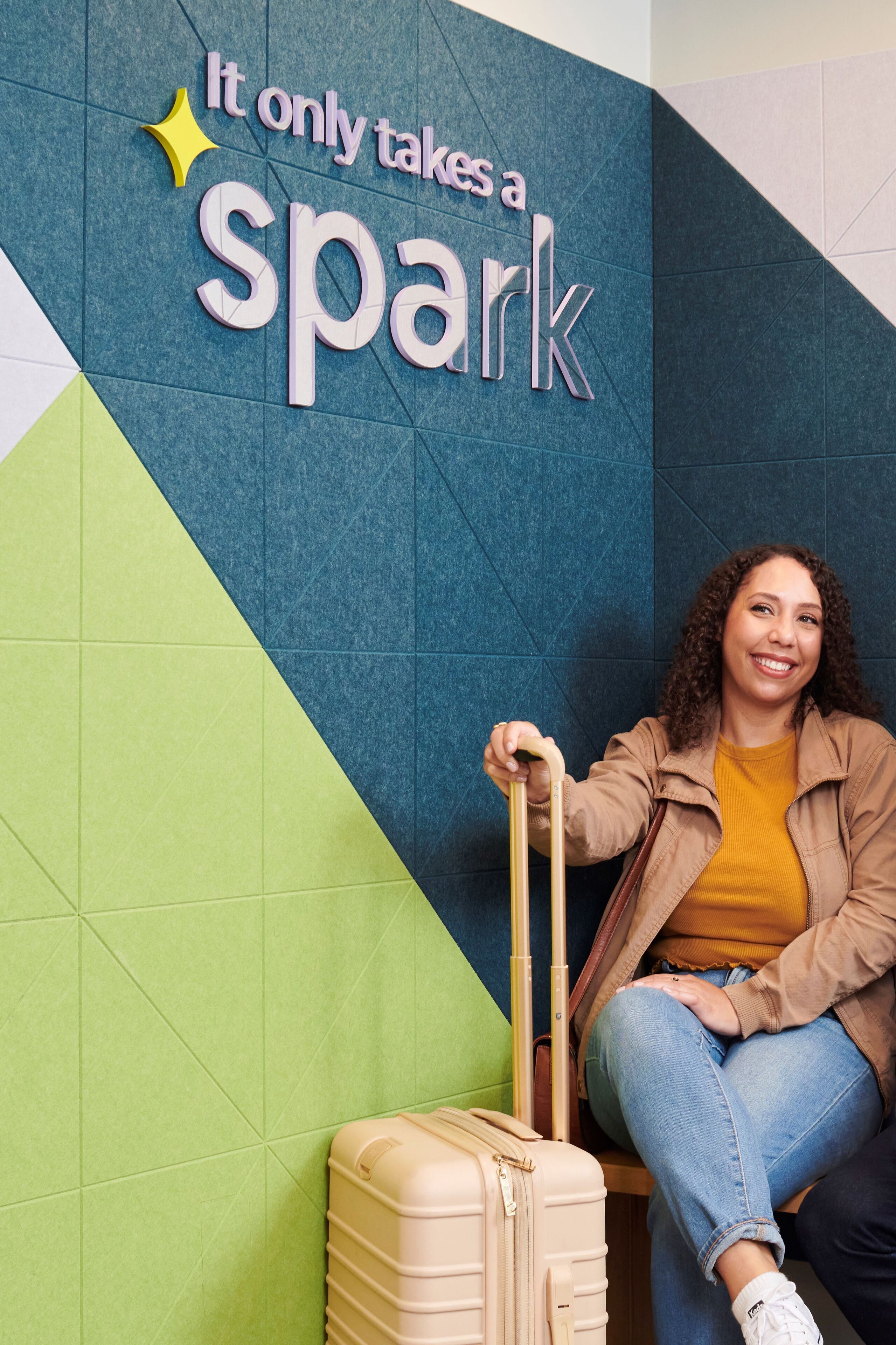 A person sitting next to a suitcase with a wall displaying the text "It only takes a spark".