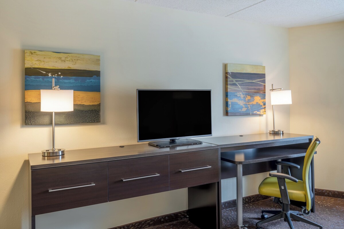A hotel room with a desk, a flat-screen TV, two wall-mounted lamps, and two paintings.