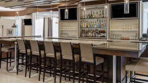 A modern bar area with a long counter, multiple stools, and a variety of bottles and glasses on shelves. - Holiday Inn Greensboro Coliseum by IHG (Greensboro)