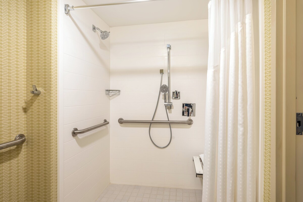A bathroom with white tiled walls, a shower with a handheld showerhead, a grab bar, and a towel rack.