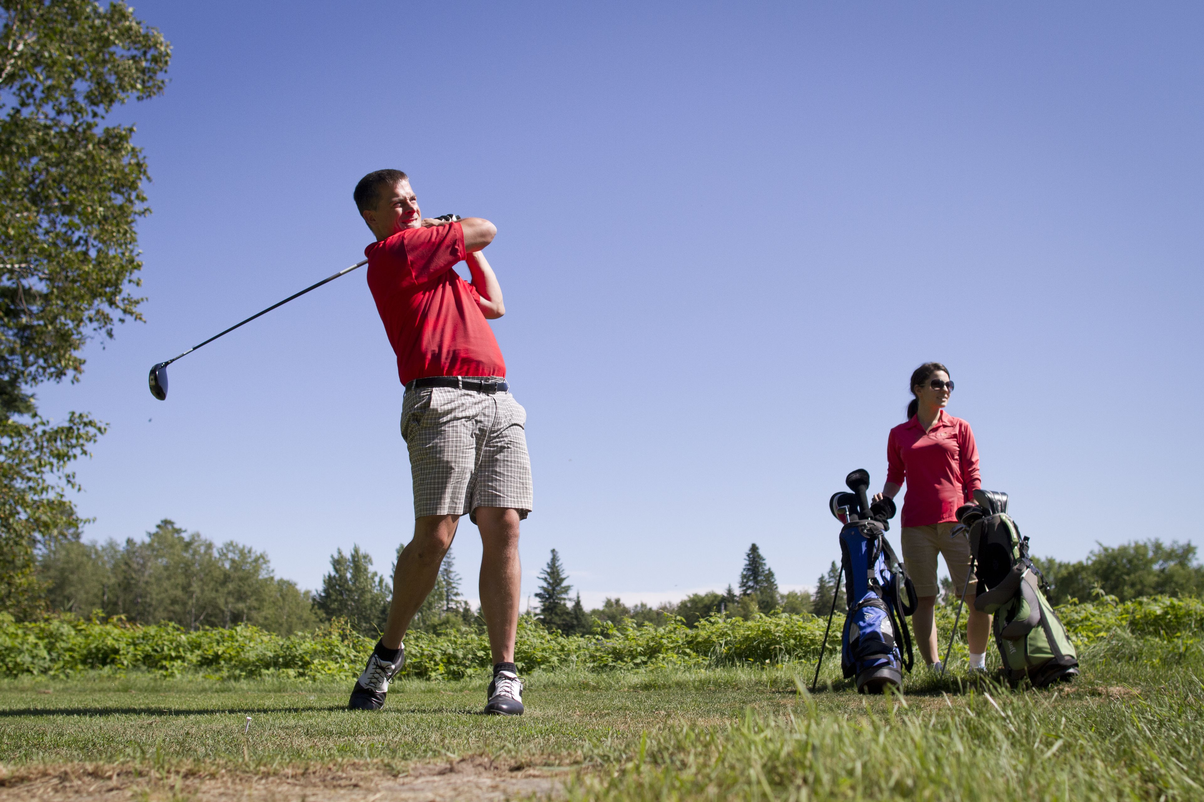 A golf course with a player preparing to swing, a woman standing nearby, and golf bags on the ground.