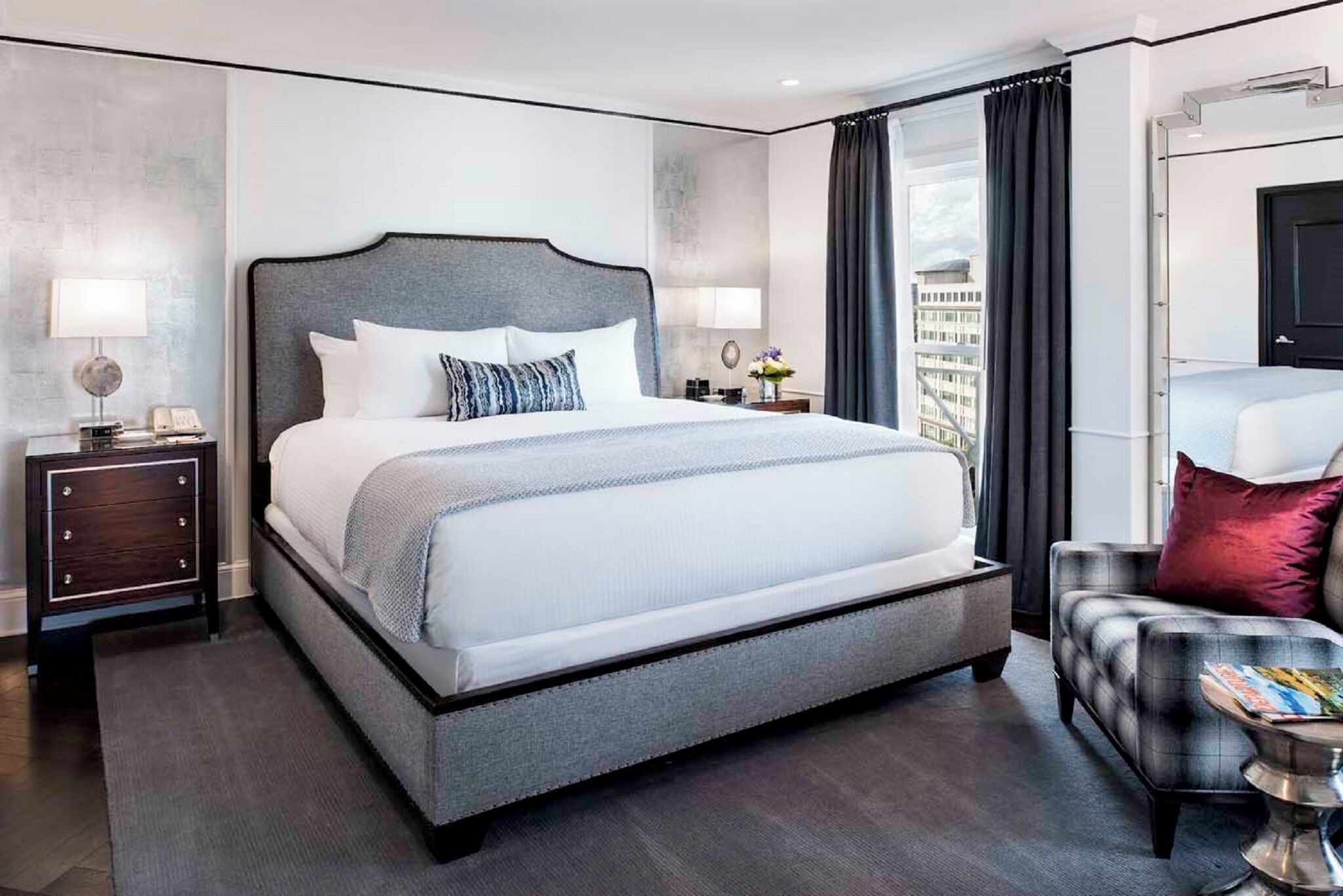 A modern hotel room with a large bed, a nightstand with a lamp, a chair, and a view of the city.