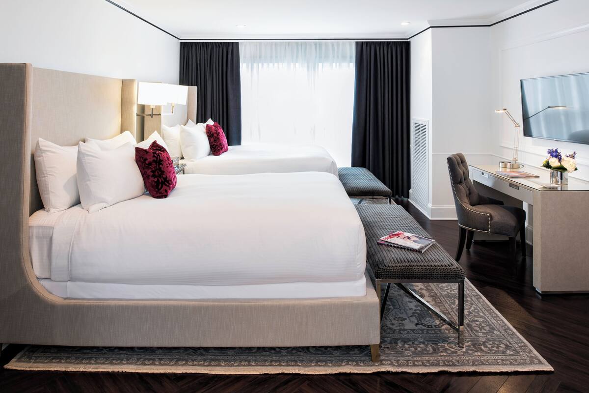 A hotel room with two beds, a desk, a chair, a TV, and a window with curtains.