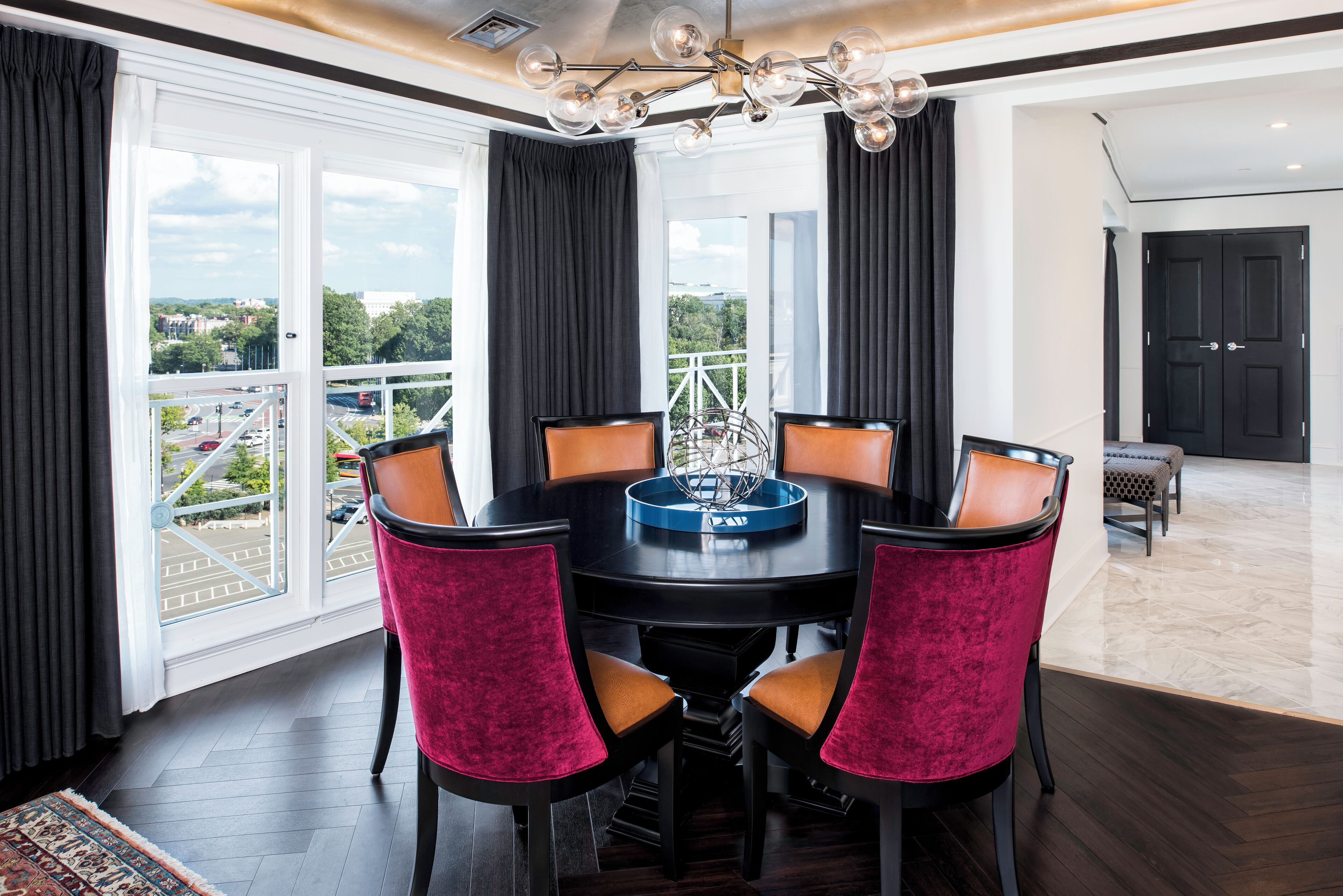 A dining area with a round table, six chairs, a chandelier, and large windows with curtains.