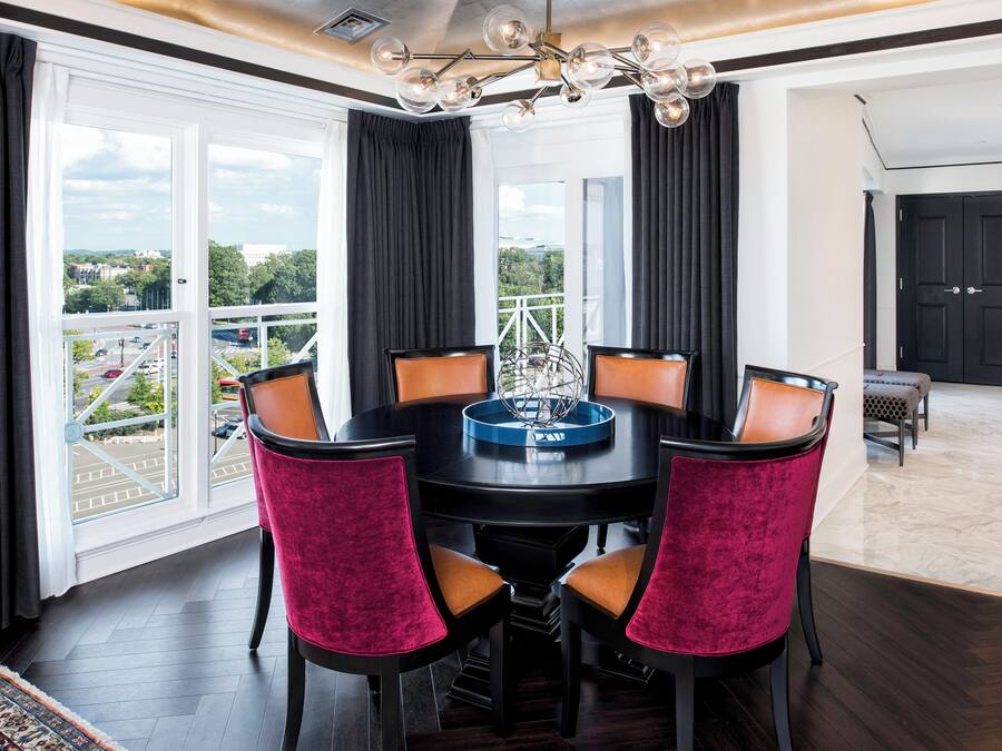 A dining area with a round table, six chairs, a chandelier, and large windows with curtains.