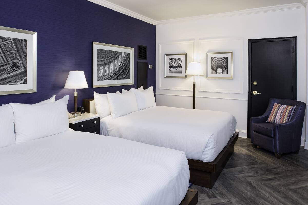 A hotel room with two beds, a nightstand, a chair, and framed pictures on the walls.