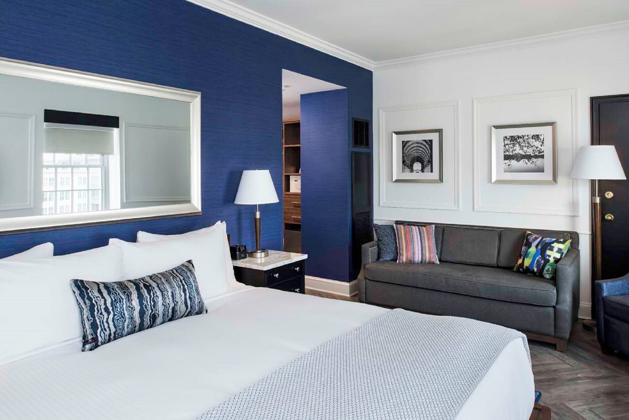 A hotel room with a large bed, a sofa, a desk with a lamp, a wardrobe, and framed pictures on the wall.